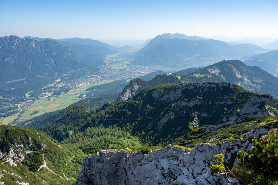 Weite Berglandschaft mit Tälern, Dörfern und einem Aussichtspunkt auf felsigem Gipfel bei klarem Himmel.