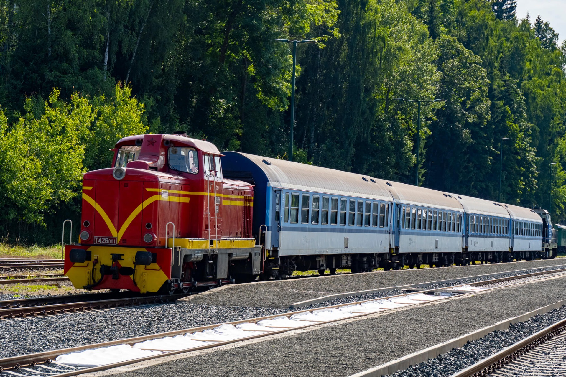 Roter Zug mit gelbem Zierstreifen und blauen Wagen steht auf Gleisen vor dichtem Wald.