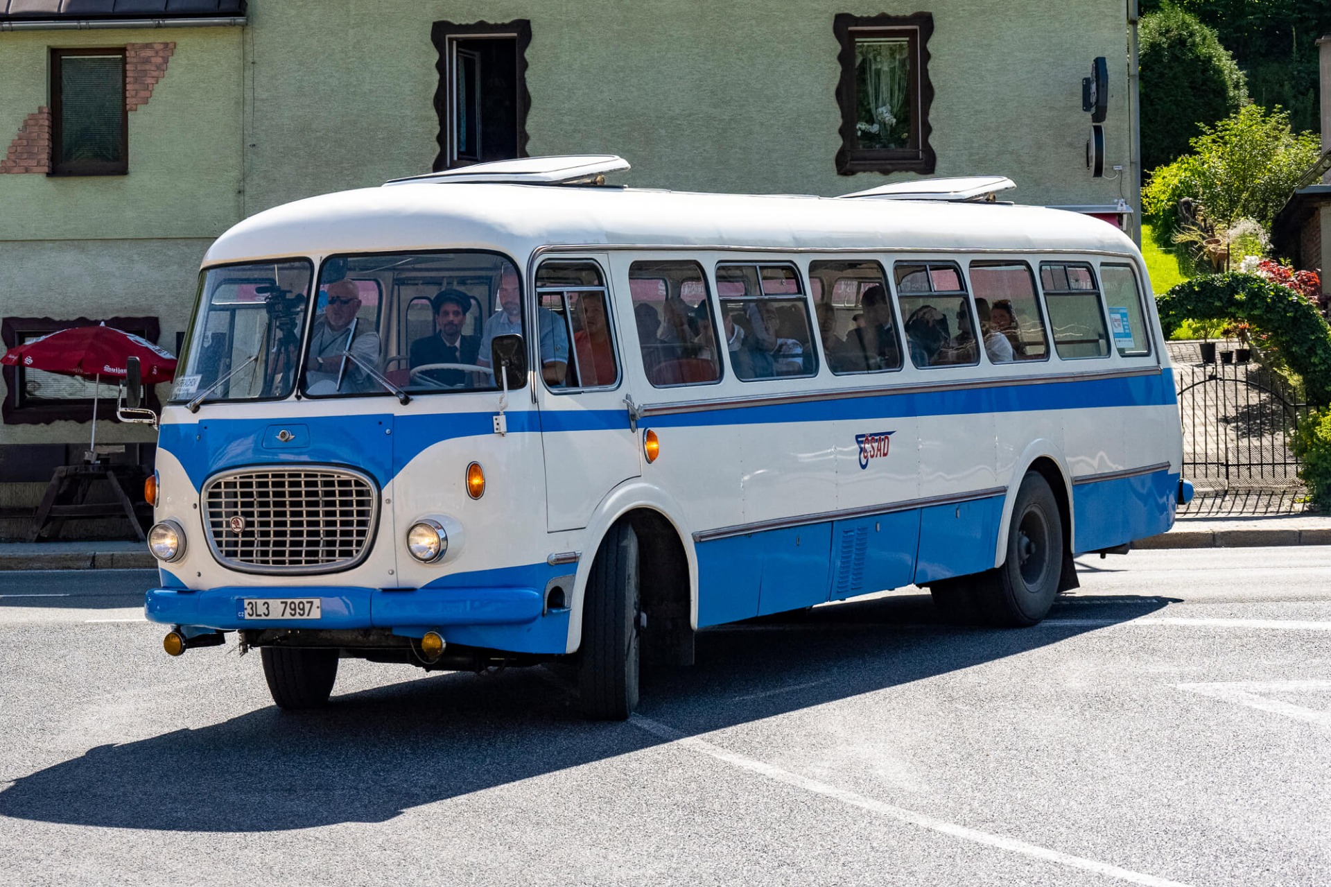 Blauer und weißer Oldtimer-Bus mit geöffneten Fenstern vor einem grünen Gebäude, Passagiere im Inneren.