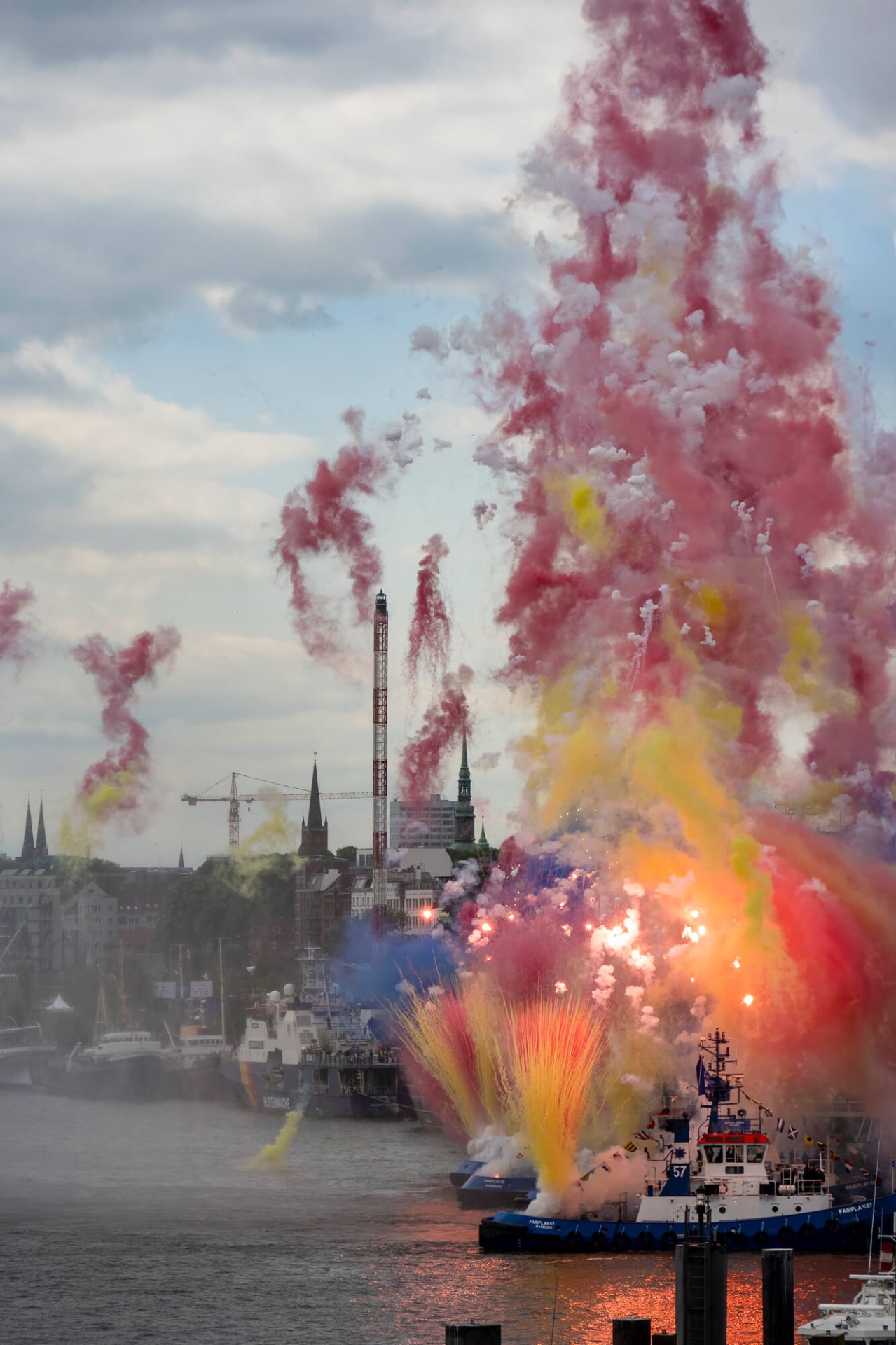 Feuerwerk mit farbigen Rauchfontänen über dem Hafen, im Hintergrund Kirchturm und Schiffe bei Abendhimmel.