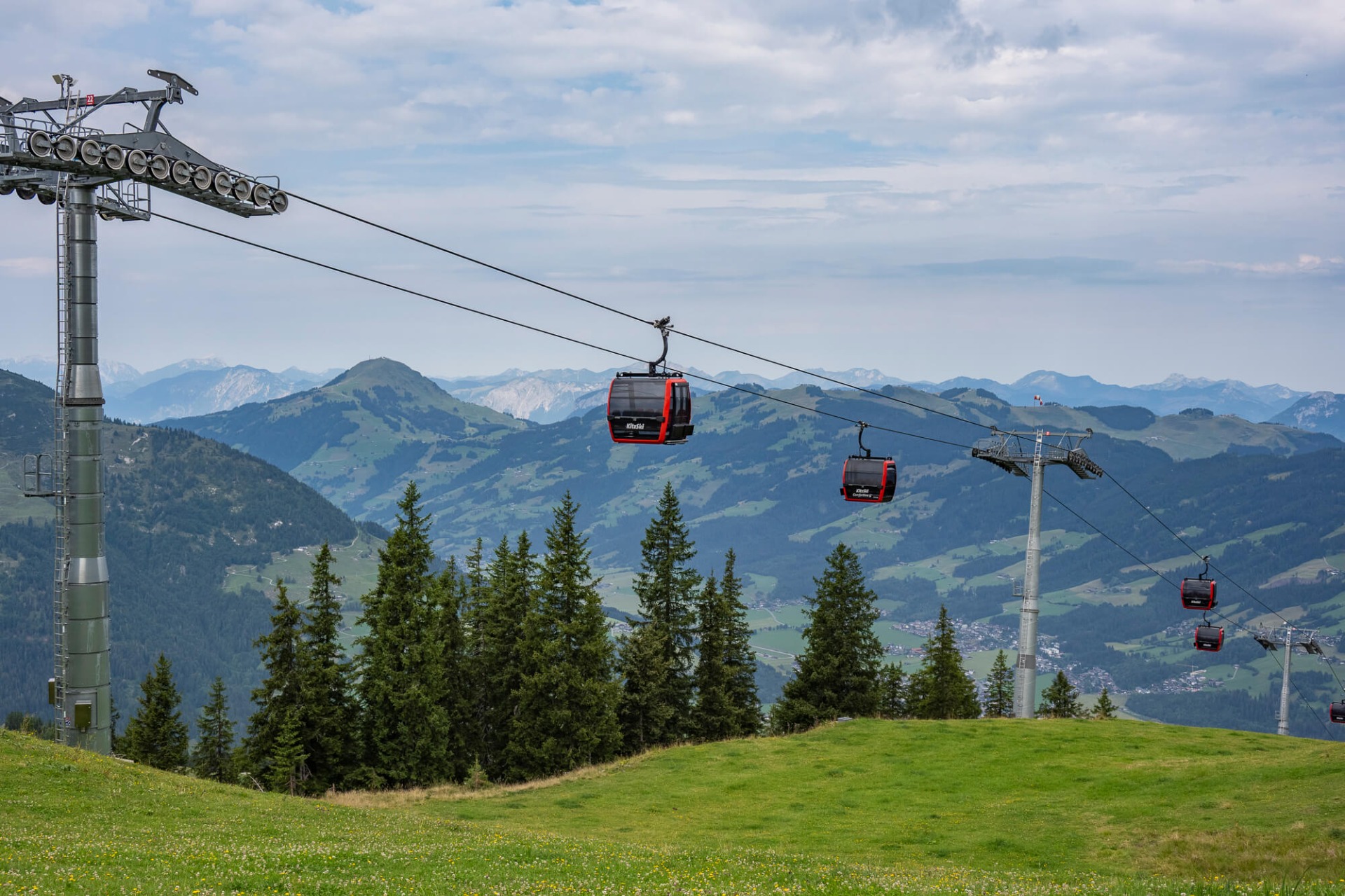 Seilbahn mit roten Kabinen über grünen Wiesen, Tannen und Berglandschaft unter bewölktem Himmel.