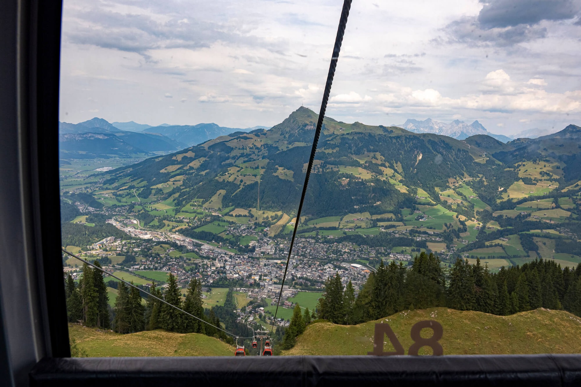 Blick aus der Gondel auf ein Tal mit Dorf, grünen Hängen und Bergen unter bewölktem Himmel.