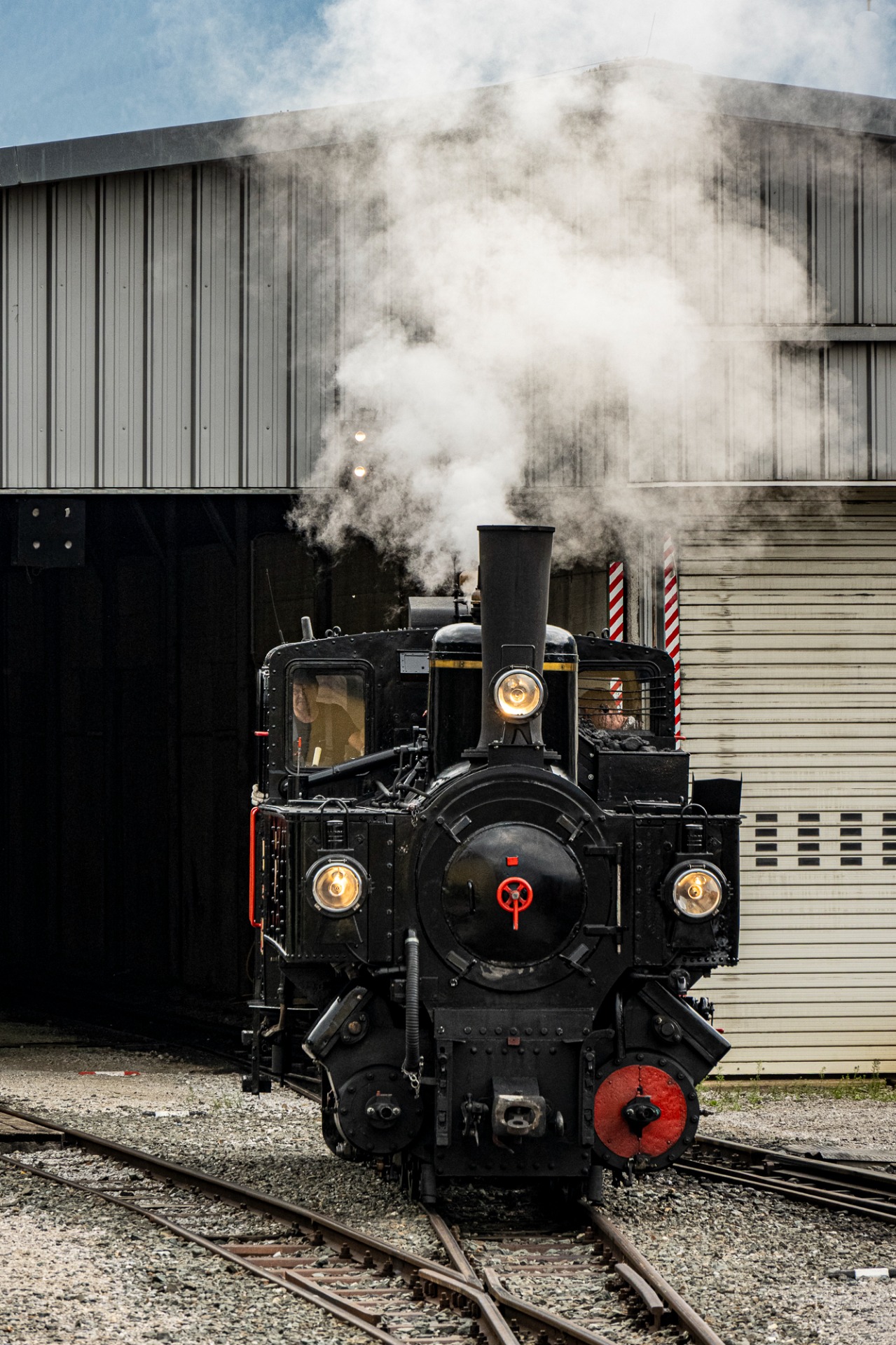 Dampfende Dampflokomotive vor Lagerhalle, Rauch steigt aus dem Schornstein, Scheinwerfer leuchten, Gleise im Vordergrund.