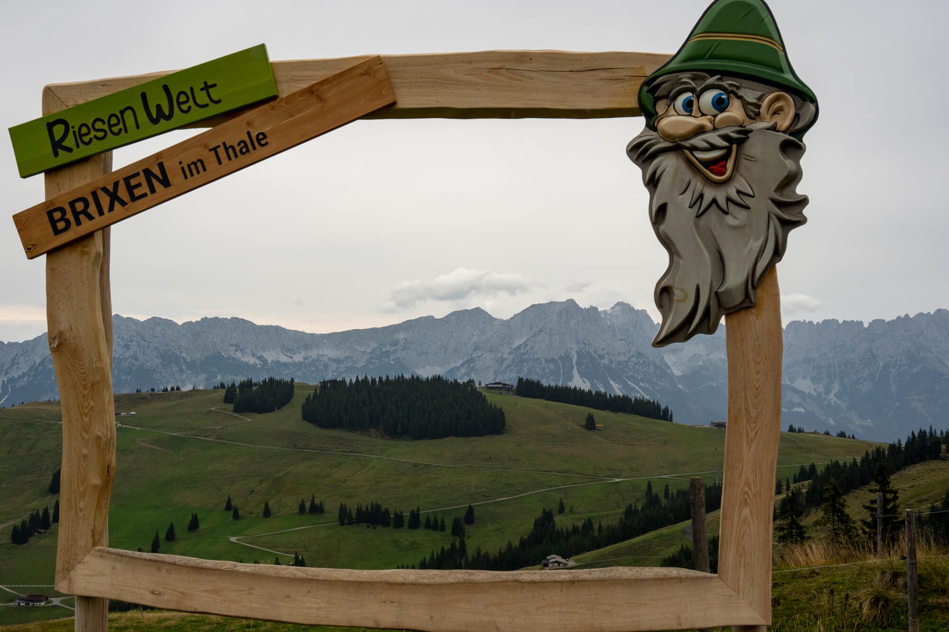 Holzbogen mit Schild „Riesenwet“ und „BRIXEN im Thale“ sowie freundlichem Bergmann im Vordergrund.