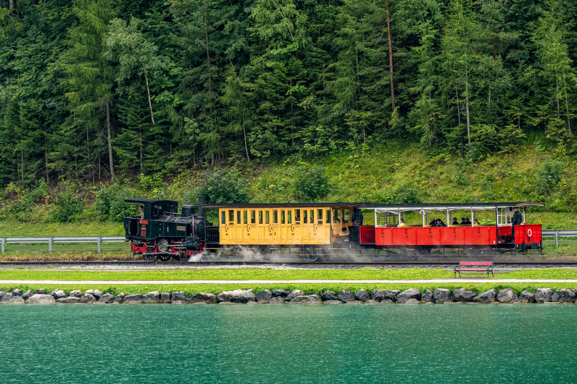 Gelber und roter Nostalgiezug fährt am See entlang, Dampf steigt auf, umgeben von dichtem Wald.