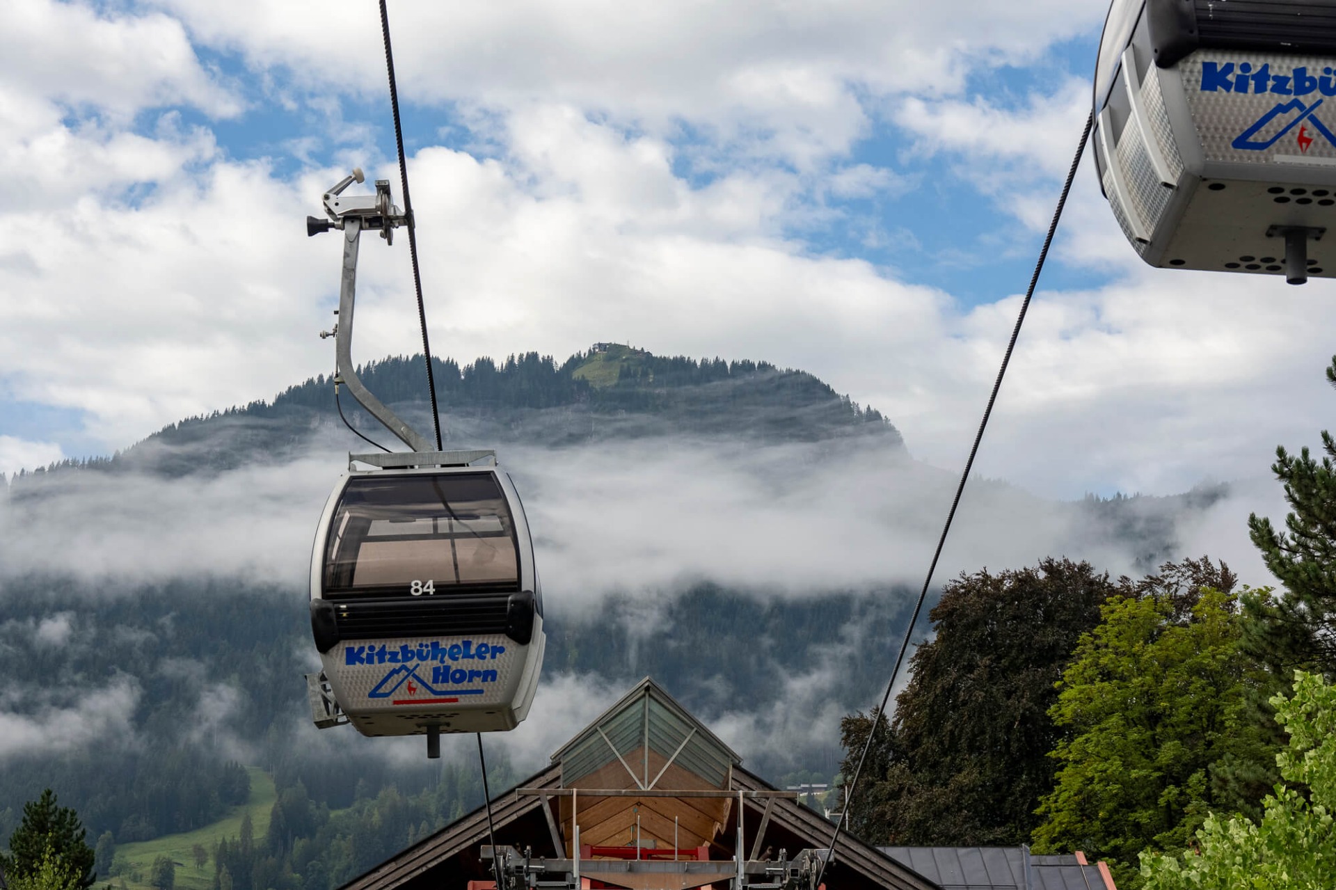 Seilbahn-Gondel über Nebel und bewaldeten Hügeln, Kabel gespannt, blauer Himmel mit Wolken.