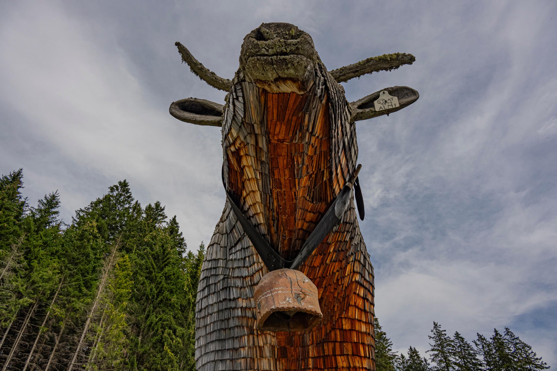 Große Holzskulptur eines Vogels mit Flügeln, Moos am Kopf, vor bewaldetem Hintergrund und wolkigem Himmel.