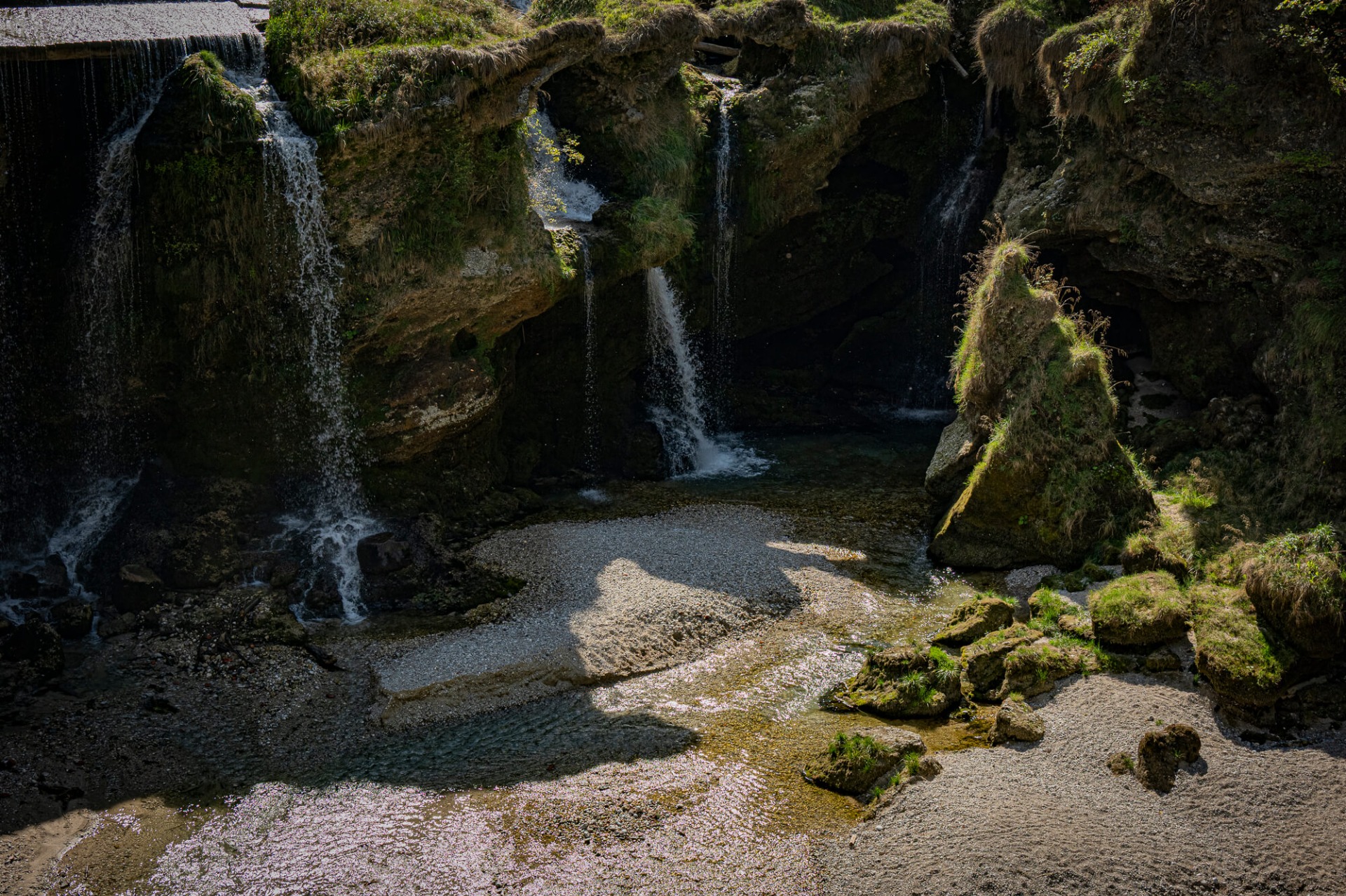 Mehrere Wasserfälle stürzen in eine felsige Schlucht, umgeben von Moos, Gras und dunklen Gesteinswänden.