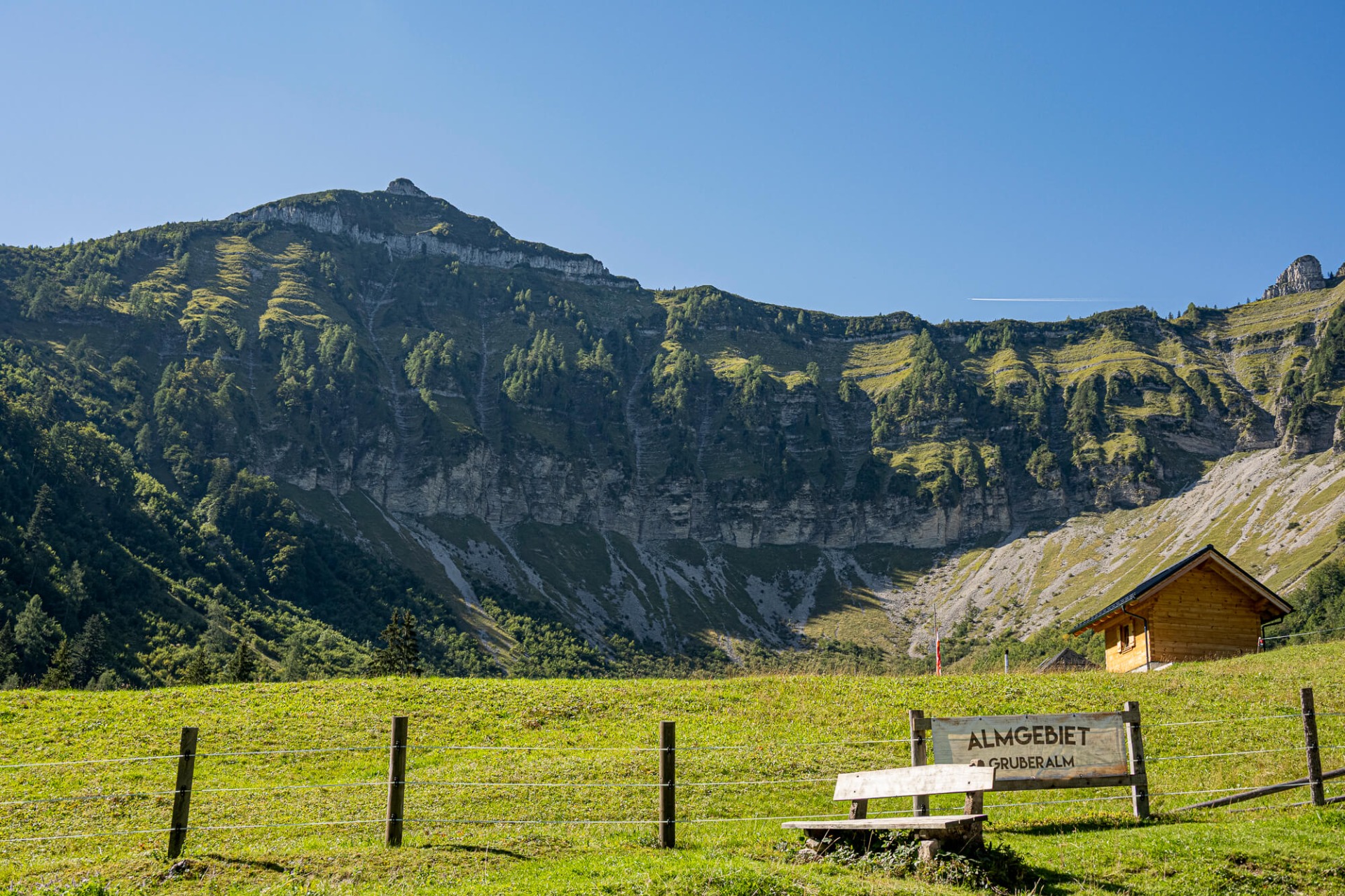 Grüne Alpwiese mit Holzhütte und Wegweiser, im Hintergrund steile Bergwand unter klarem blauen Himmel.