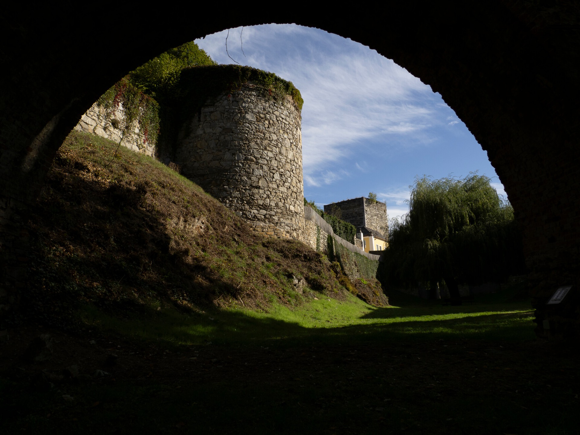 Blick durch einen dunklen Torbogen auf eine alte Steinmauer, bewachsenem Hügel und grünen Garten.