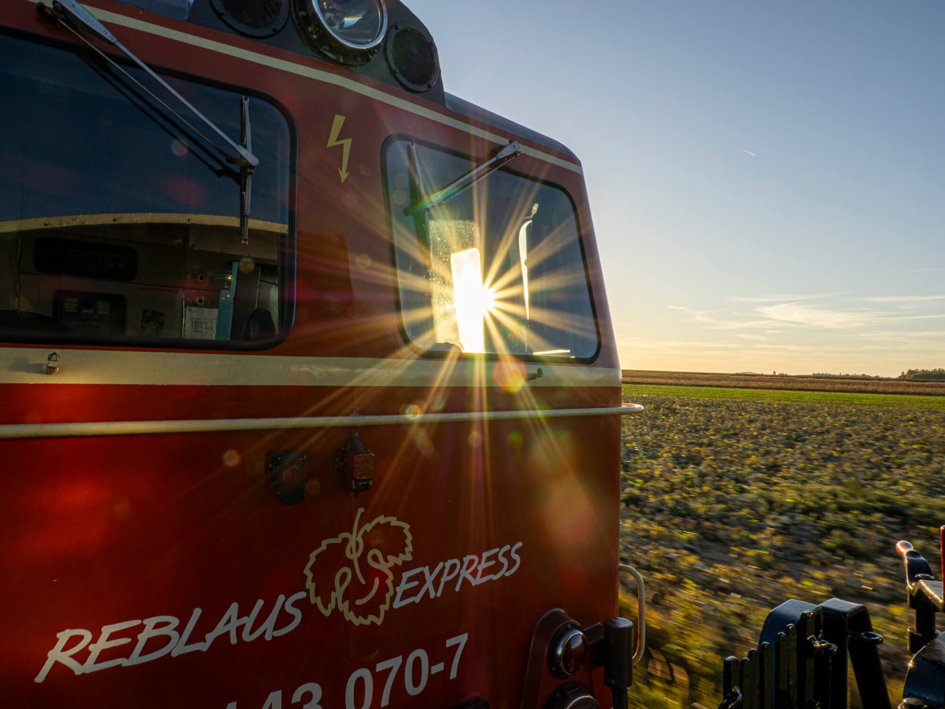 Roter Zug mit geöffnetem Fenster, Funken und Sonnenstrahlen, vorbeifahrend über Felder bei Sonnenuntergang.