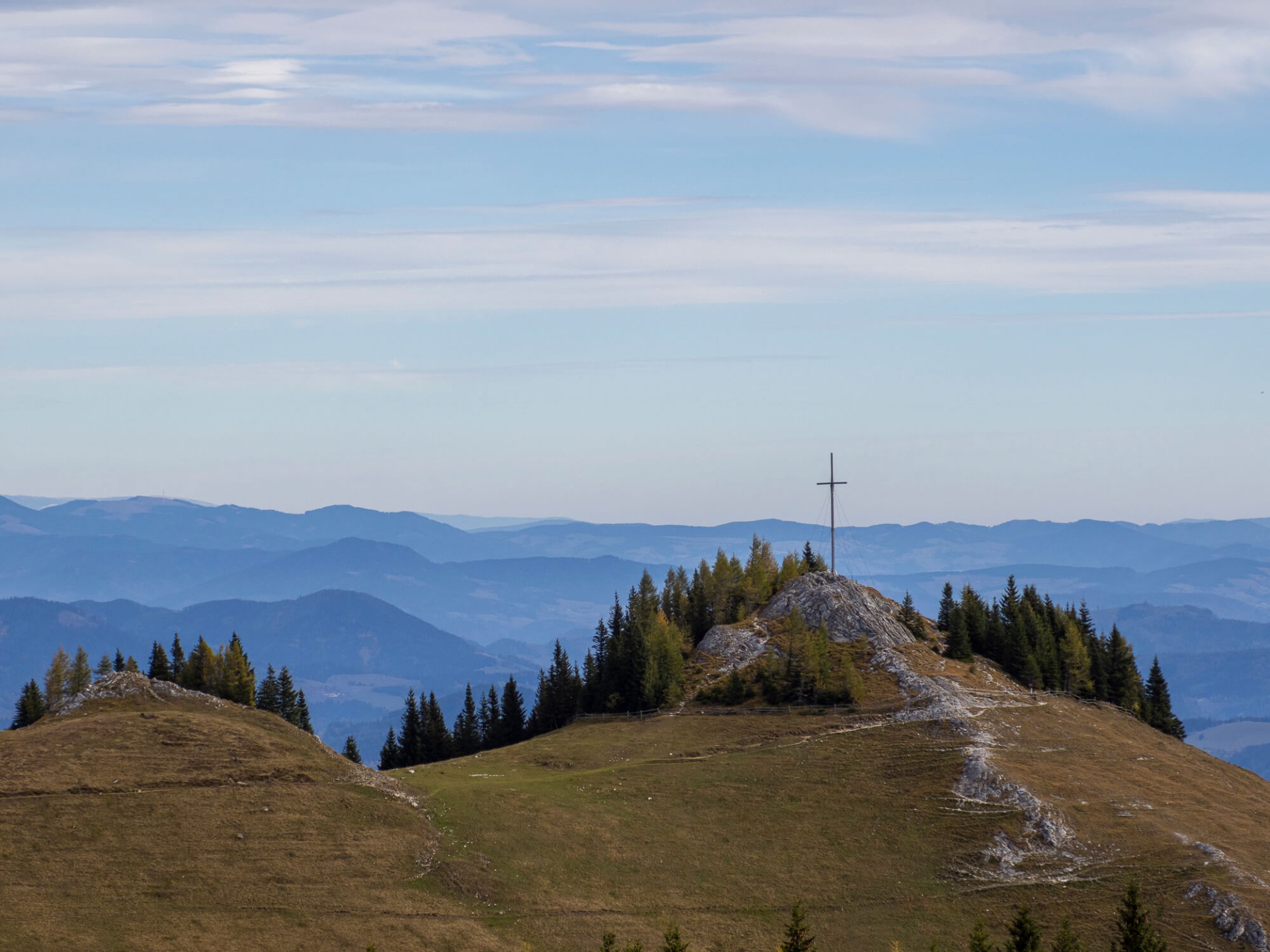 Berggipfel mit Kreuz auf Fels, Tannenwald und weiten Hügeln unter blauem, wolkigem Himmel.